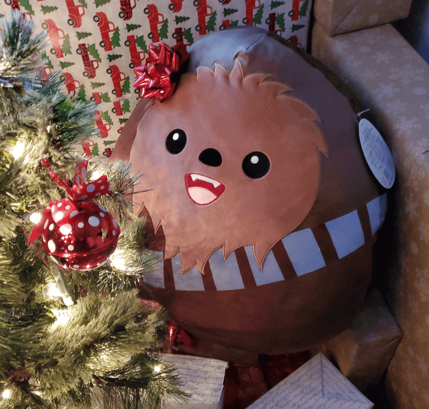 A cute plush toy resembling a Christmas-themed character with a bow, smiling near a decorated tree.